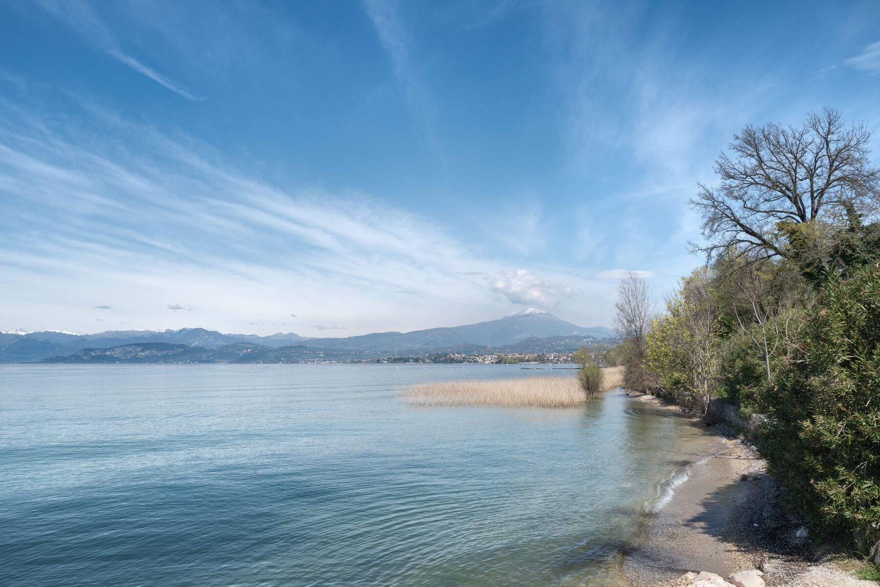 Naturbelassenes Kiesufer am Gardasee bei Lazise mit Schilf, klarem Wasser und Bergkulisse